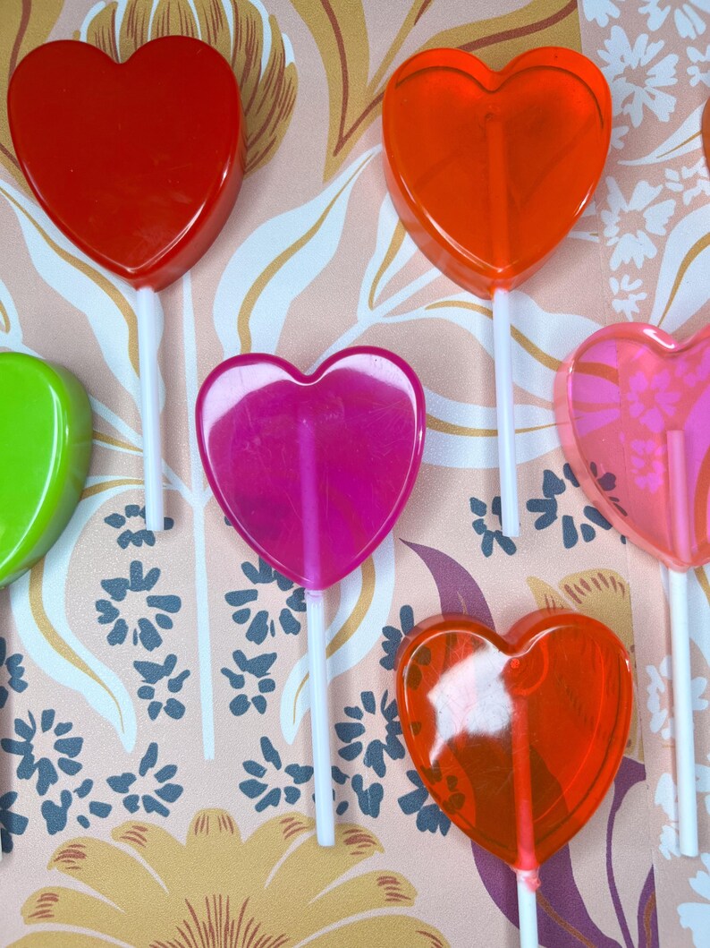 May include: A selection of heart-shaped lollipops in shades of red, orange, pink, and green, each on a white stick. The lollipops are displayed against a patterned backdrop with floral and leaf designs.