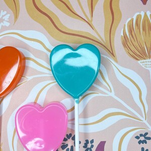 May include: Three heart-shaped lollipops in orange, teal, and pink. Each lollipop is on a white stick. The background has a floral pattern with pink, yellow, and purple tones.