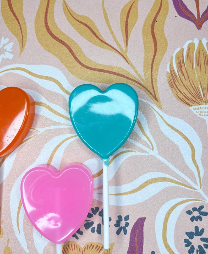 May include: Three heart-shaped lollipops in orange, teal, and pink. Each lollipop is on a white stick. The background has a floral pattern with pink, yellow, and purple tones.