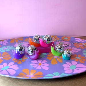 May include: A collection of disco ball snail pendants in various colors, including purple, pink, orange, green, and turquoise, displayed on a floral patterned tray. Each snail has a silver chain.