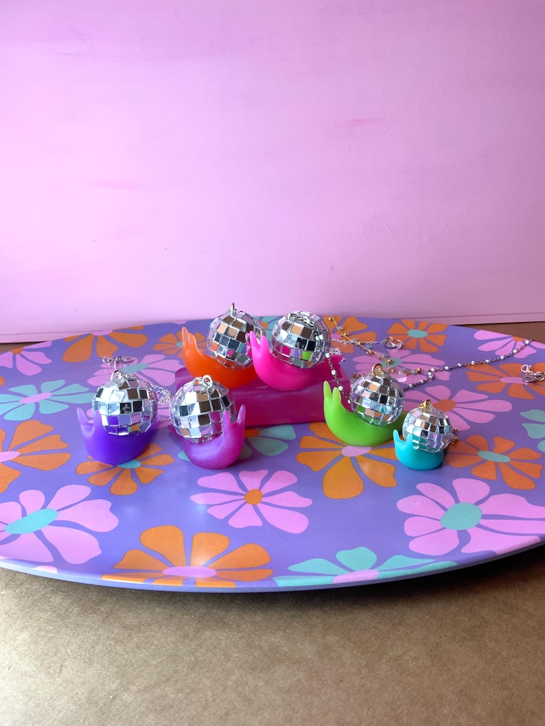 May include: A collection of disco ball snail pendants in various colors, including purple, pink, orange, green, and turquoise, displayed on a floral patterned tray. Each snail has a silver chain.