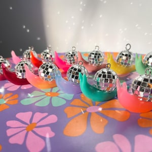 May include: A collection of colorful, snail-shaped ornaments, each topped with a miniature disco ball. The ornaments are arranged on a purple surface with a floral pattern. The disco balls reflect light, creating a sparkling effect.