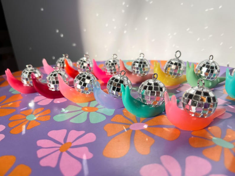 May include: A collection of colorful, snail-shaped ornaments, each topped with a miniature disco ball. The ornaments are arranged on a purple surface with a floral pattern. The disco balls reflect light, creating a sparkling effect.