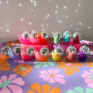 May include: A collection of colorful snail-shaped ornaments, each topped with a miniature disco ball. The snails are in various bright colors, including pink, orange, yellow, green, blue, and purple, arranged on a floral patterned surface.