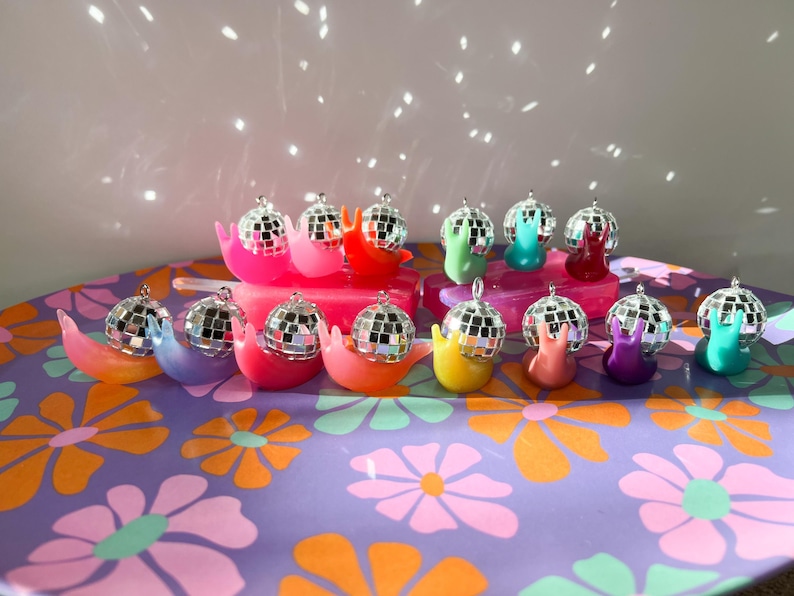 May include: A collection of colorful snail-shaped ornaments, each topped with a miniature disco ball. The snails are in various bright colors, including pink, orange, yellow, green, blue, and purple, arranged on a floral patterned surface.