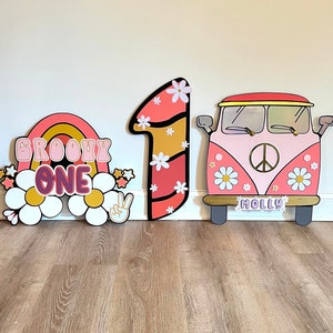 May include: Three colorful wooden decorations for a groovy themed party. The decorations include a rainbow with the words "Groovy One", a pink and orange number one, and a pink and white Volkswagen bus with the word "Molly" on the front.