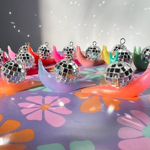 May include: A collection of disco ball ornaments atop colorful, curved, snail-shaped holders. The holders are in shades of pink, orange, purple, green, and blue, arranged on a floral patterned surface. The disco balls reflect light.