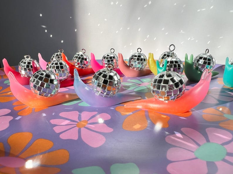 May include: A collection of disco ball ornaments atop colorful, curved, snail-shaped holders. The holders are in shades of pink, orange, purple, green, and blue, arranged on a floral patterned surface. The disco balls reflect light.