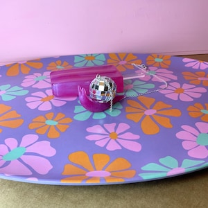 May include: A purple serving tray with a floral pattern in orange, pink, and teal. A pink popsicle-shaped object and a disco ball necklace rest on the tray. The background is a solid pink.