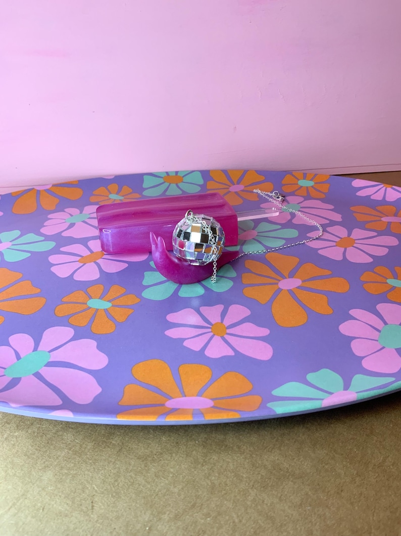 May include: A purple serving tray with a floral pattern in orange, pink, and teal. A pink popsicle-shaped object and a disco ball necklace rest on the tray. The background is a solid pink.