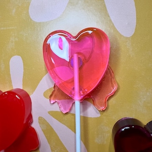 May include: A close-up of several heart-shaped lollipops on white sticks. The lollipops are various shades of red and pink, with a glossy, transparent appearance. The background is a textured yellow surface with white floral shapes.
