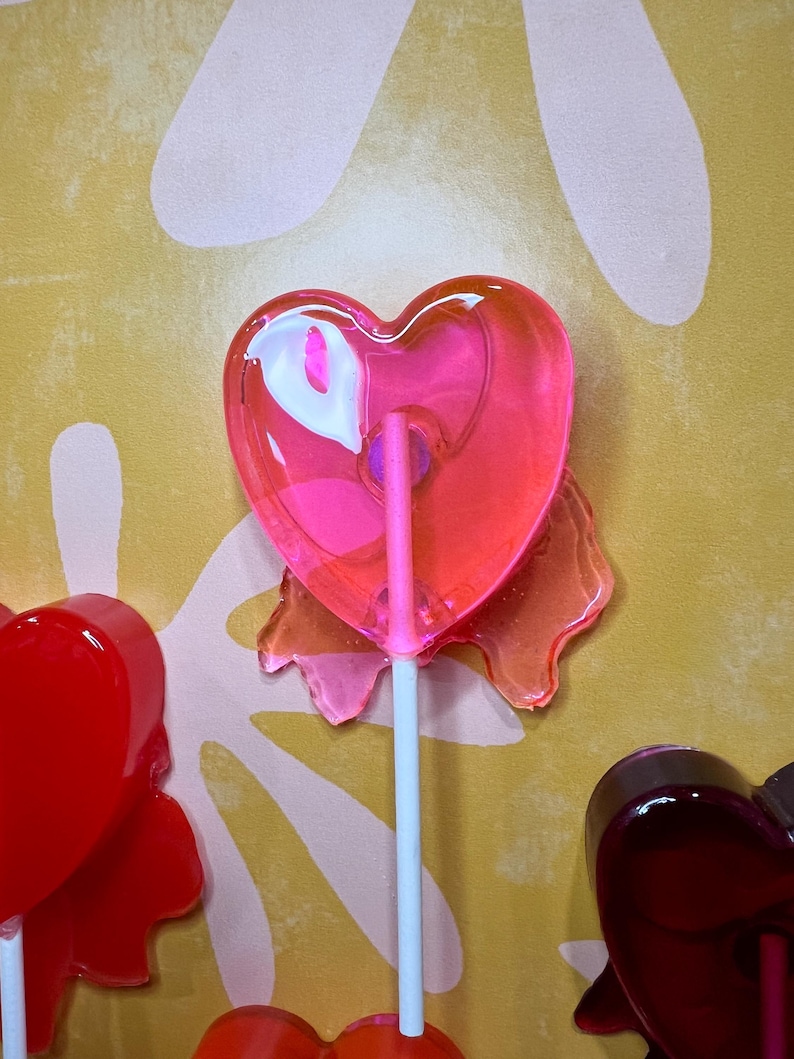 May include: A close-up of several heart-shaped lollipops on white sticks. The lollipops are various shades of red and pink, with a glossy, transparent appearance. The background is a textured yellow surface with white floral shapes.