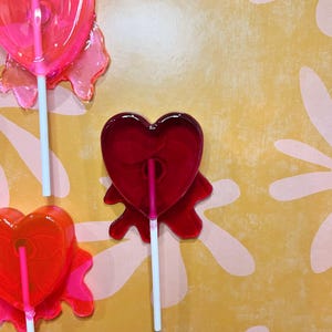 May include: Close-up of three heart-shaped lollipops on white sticks. The lollipops are various shades of red and pink, with a clear, glossy appearance. The background is a yellow surface with a pink floral pattern.