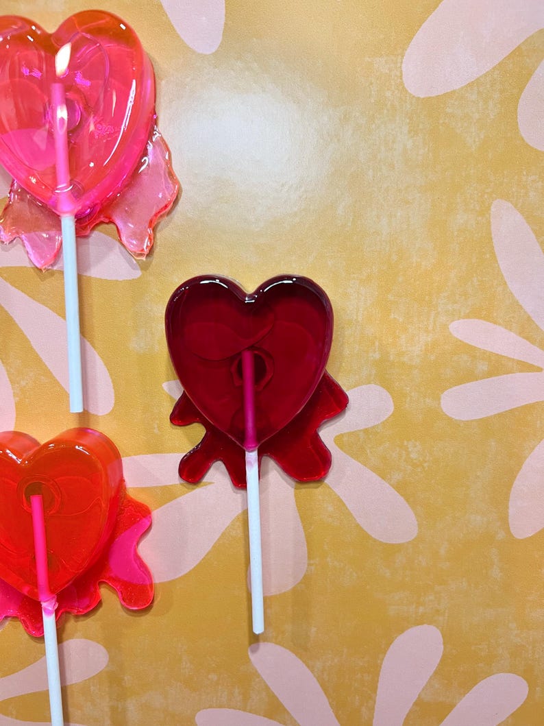 May include: Close-up of three heart-shaped lollipops on white sticks. The lollipops are various shades of red and pink, with a clear, glossy appearance. The background is a yellow surface with a pink floral pattern.