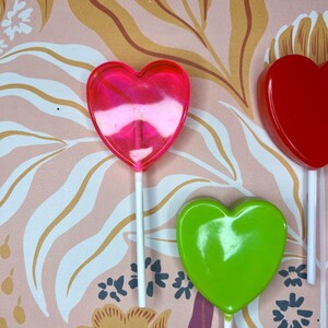 May include: Image of heart-shaped lollipops in pink, red, and green. Each lollipop is on a white stick. The background features a pink floral pattern with gold and white accents.