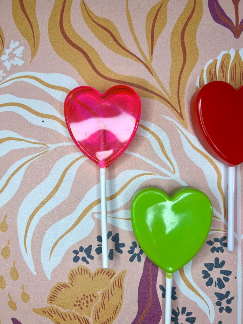 May include: Image of heart-shaped lollipops in pink, red, and green. Each lollipop is on a white stick. The background features a pink floral pattern with gold and white accents.