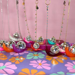 May include: A collection of necklaces with disco ball pendants displayed on colorful, snail-shaped holders. The necklaces hang against a pink backdrop, resting on a tray with a floral pattern. The holders are in various colors.