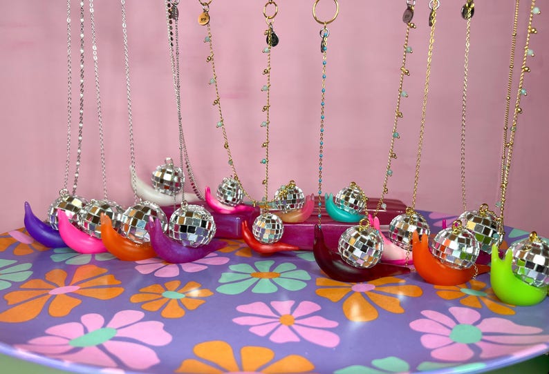 May include: A collection of necklaces with disco ball pendants displayed on colorful, snail-shaped holders. The necklaces hang against a pink backdrop, resting on a tray with a floral pattern. The holders are in various colors.