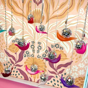 May include: A collection of decorative hanging ornaments. Each ornament features a disco ball atop a colorful snail figurine. The snails are in various colors, including red, pink, orange, and purple, suspended from chains against a floral wallpaper backdrop.
