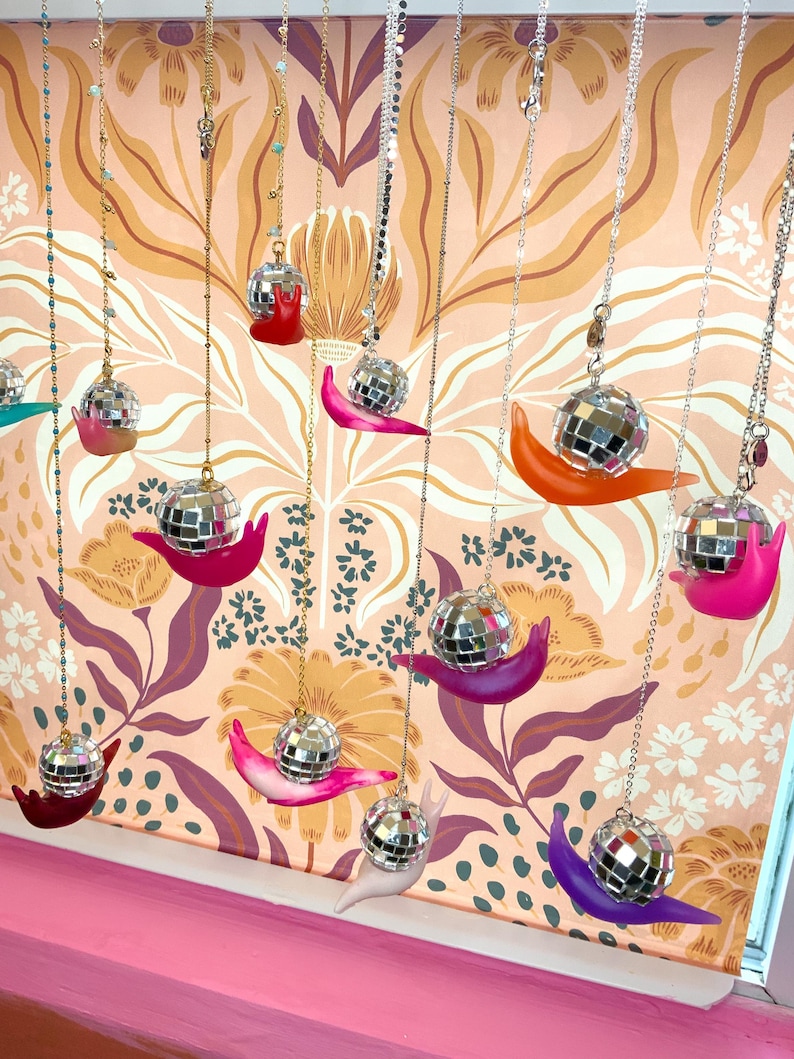May include: A collection of decorative hanging ornaments. Each ornament features a disco ball atop a colorful snail figurine. The snails are in various colors, including red, pink, orange, and purple, suspended from chains against a floral wallpaper backdrop.