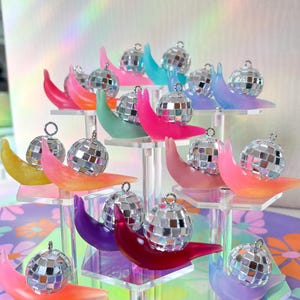 May include: A display of miniature disco ball ornaments, each perched on a colorful, crescent-shaped base. The bases are in shades of pink, orange, yellow, green, blue, and purple, arranged on a clear, tiered stand. The disco balls are silver and reflective.