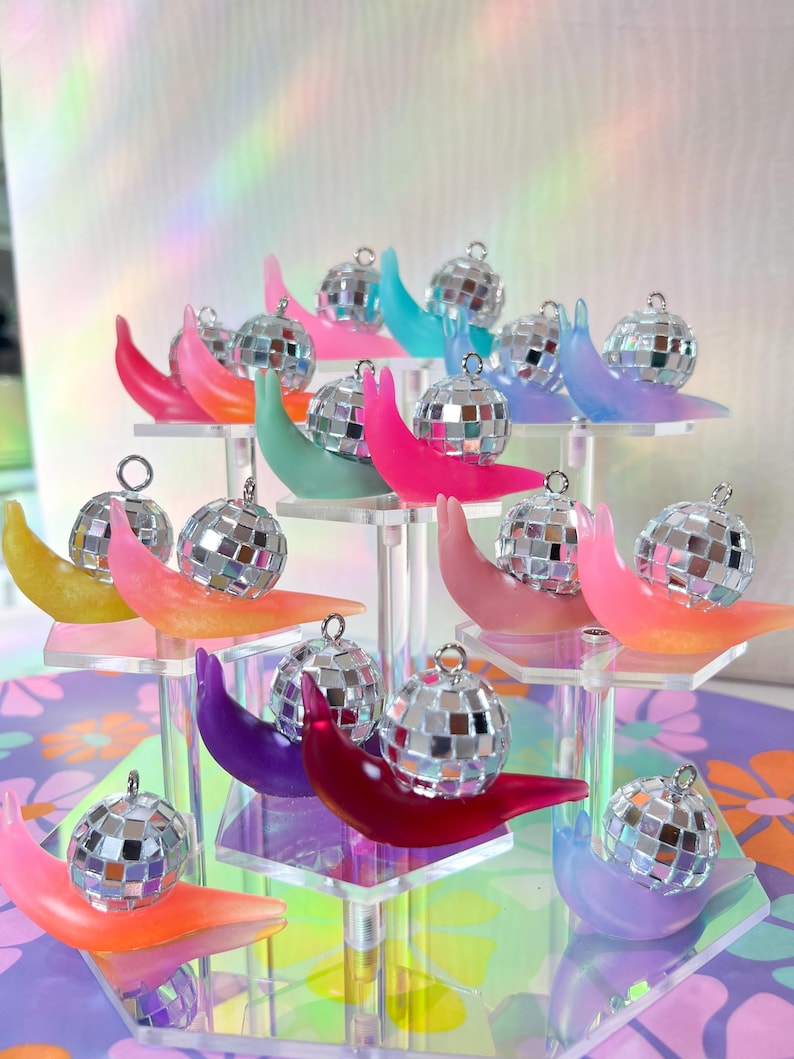 May include: A display of miniature disco ball ornaments, each perched on a colorful, crescent-shaped base. The bases are in shades of pink, orange, yellow, green, blue, and purple, arranged on a clear, tiered stand. The disco balls are silver and reflective.