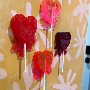 May include: Four heart-shaped lollipops in shades of red and pink are displayed against a yellow and pink patterned background. Each lollipop is on a white stick and has a translucent appearance, with a ribbon-like detail at the base of the heart.