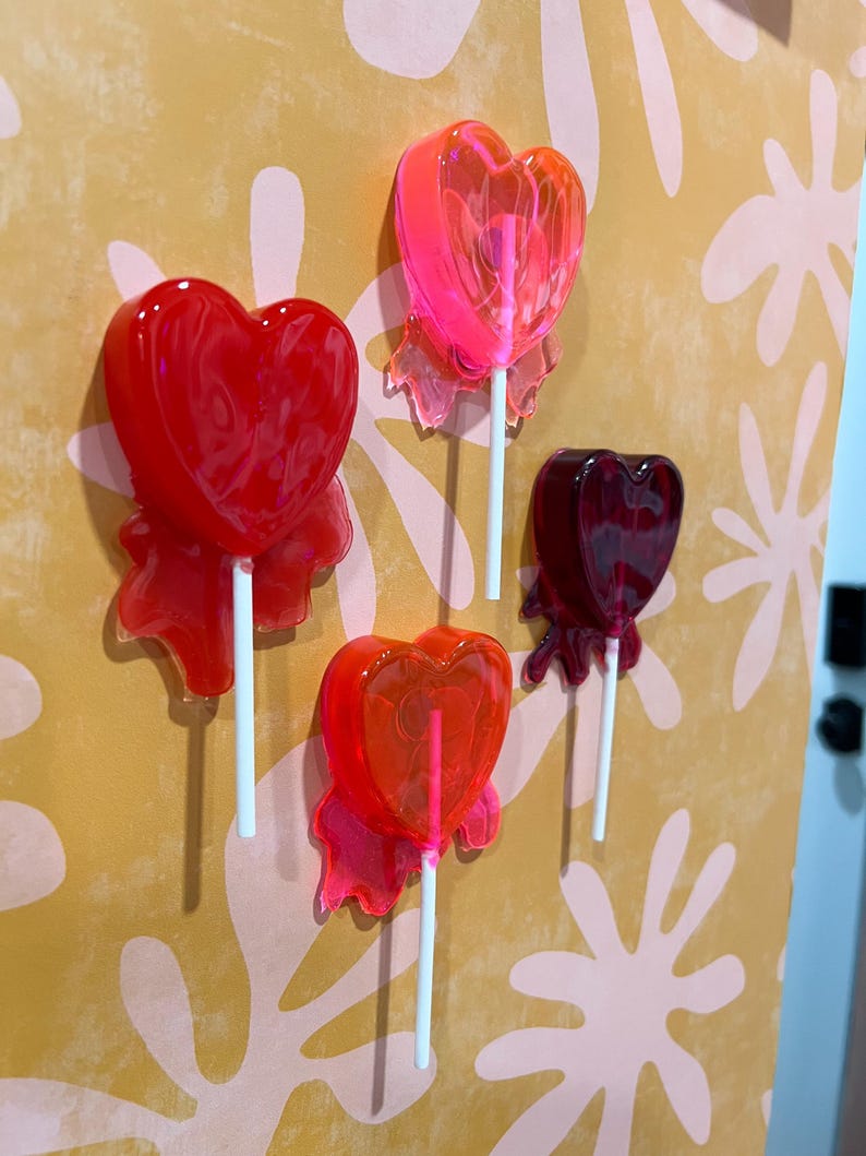May include: Four heart-shaped lollipops in shades of red and pink are displayed against a yellow and pink patterned background. Each lollipop is on a white stick and has a translucent appearance, with a ribbon-like detail at the base of the heart.