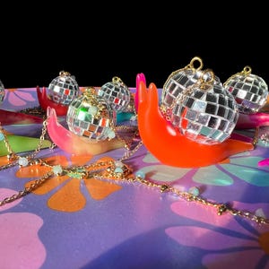 May include: A collection of disco ball ornaments atop colorful, snail-shaped sculptures. The ornaments are silver and reflective, with gold chains and small, light blue beads. The sculptures are in shades of orange, pink, and green, set against a floral patterned background.