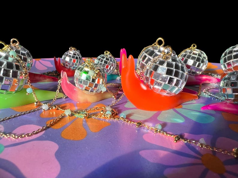 May include: A collection of disco ball ornaments atop colorful, snail-shaped sculptures. The ornaments are silver and reflective, with gold chains and small, light blue beads. The sculptures are in shades of orange, pink, and green, set against a floral patterned background.