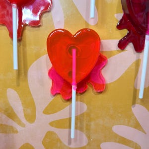 May include: Close-up of several heart-shaped lollipops on white sticks. The lollipops are a vibrant red color, with a glossy, transparent appearance. The background is a textured yellow surface with pink floral patterns.