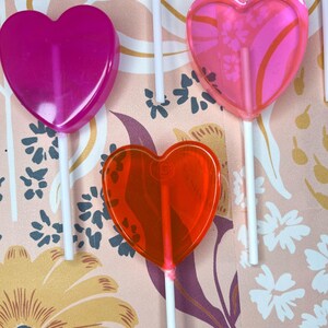 May include: A selection of heart-shaped lollipops in pink and red hues, each on a white stick. The background features a floral pattern with pink, yellow, and purple flowers, creating a colourful display.