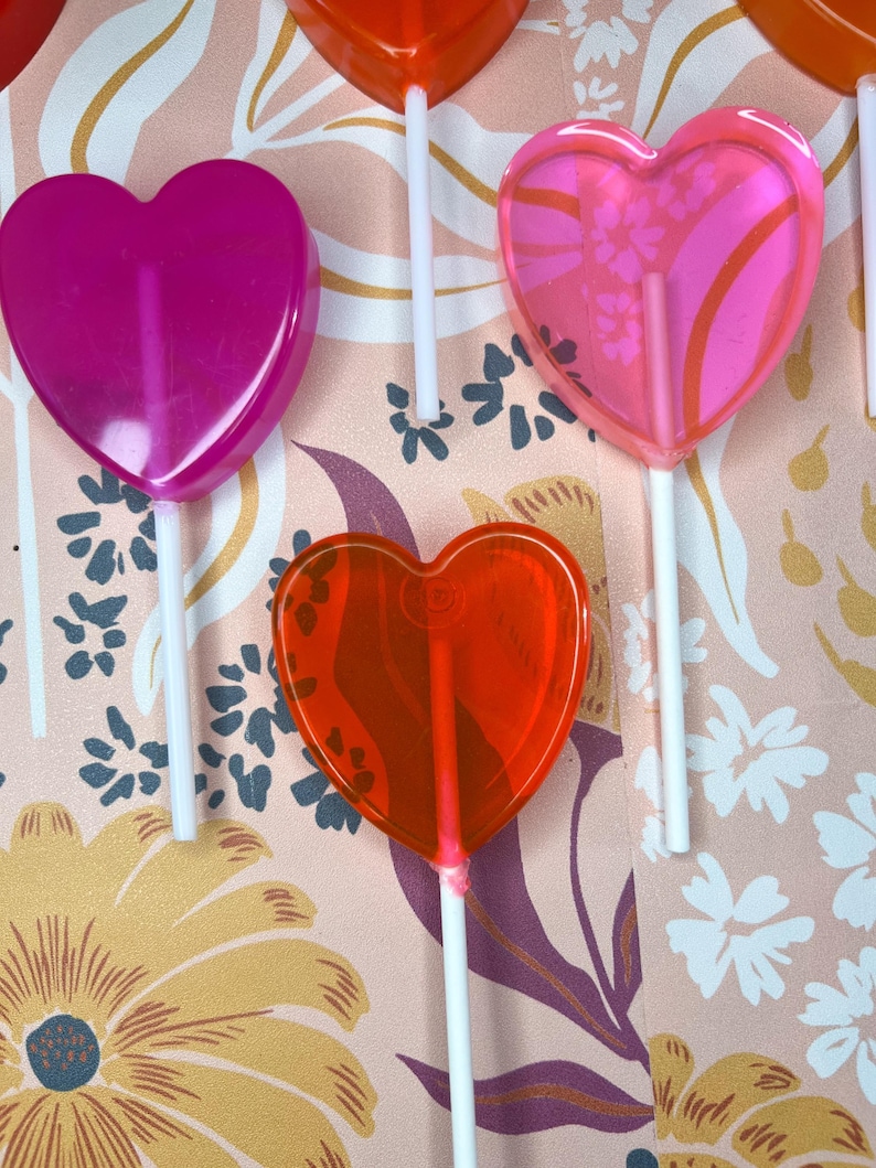 May include: A selection of heart-shaped lollipops in pink and red hues, each on a white stick. The background features a floral pattern with pink, yellow, and purple flowers, creating a colourful display.