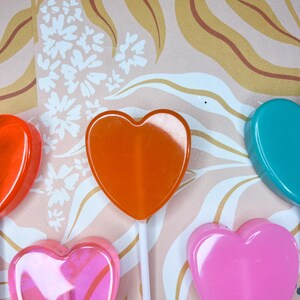 May include: A selection of heart-shaped lollipops in colours such as orange, pink, and teal, on white sticks. The lollipops are set against a patterned backdrop with floral and leaf designs.