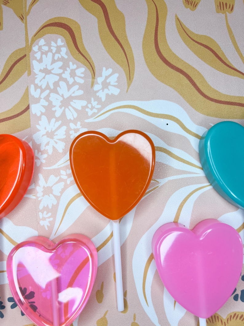 May include: A selection of heart-shaped lollipops in colours such as orange, pink, and teal, on white sticks. The lollipops are set against a patterned backdrop with floral and leaf designs.