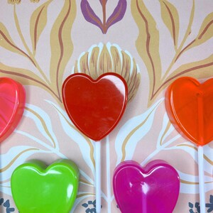 May include: A selection of colourful heart-shaped lollipops in red, orange, pink, and green, each on a white stick. The background displays a floral pattern in pink, yellow, and purple hues.