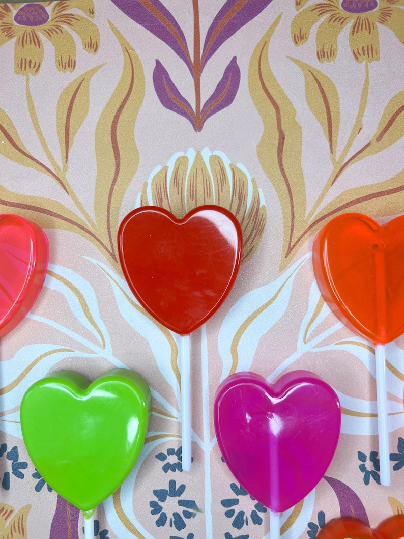 May include: A selection of colourful heart-shaped lollipops in red, orange, pink, and green, each on a white stick. The background displays a floral pattern in pink, yellow, and purple hues.