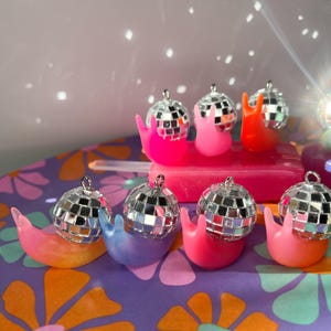 May include: A collection of colorful snail figurines, each topped with a miniature disco ball. The snails are in various bright colors, including pink, orange, blue, and green. The background features a floral pattern and a light effect.