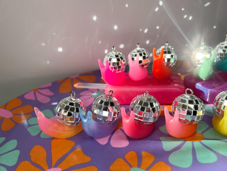 May include: A collection of colorful snail figurines, each topped with a miniature disco ball. The snails are in various bright colors, including pink, orange, blue, and green. The background features a floral pattern and a light effect.