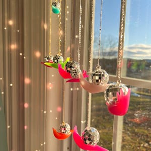 May include: A collection of disco ball pendants hanging in front of a window. Each pendant features a small mirrored ball suspended from a delicate chain, resting on a crescent-shaped base in various bright colors, including pink, orange, and green.