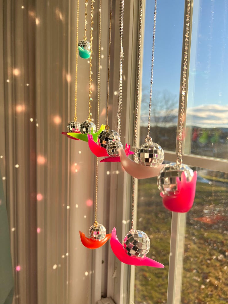 May include: A collection of disco ball pendants hanging in front of a window. Each pendant features a small mirrored ball suspended from a delicate chain, resting on a crescent-shaped base in various bright colors, including pink, orange, and green.