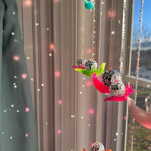 May include: Decorative hanging disco ball ornaments. Each ornament features a mirrored disco ball suspended from a gold chain, resting on a colorful, curved base. The bases are in shades of green, pink, and orange. The background includes a window and a curtain.