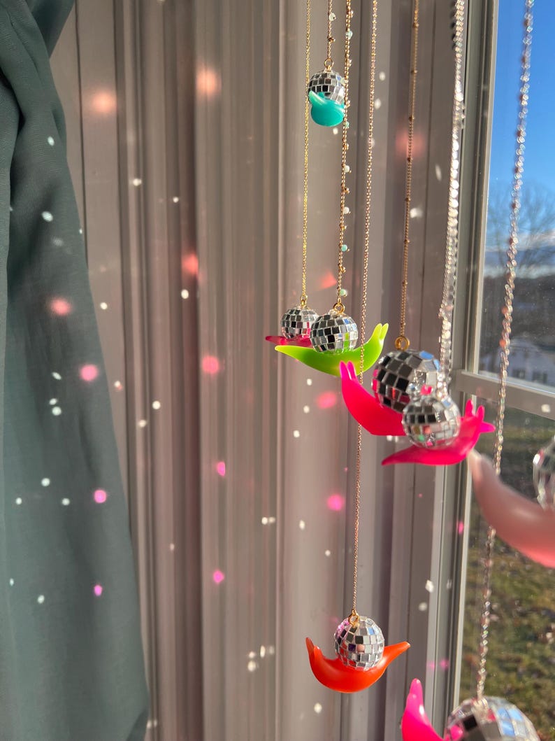 May include: Decorative hanging disco ball ornaments. Each ornament features a mirrored disco ball suspended from a gold chain, resting on a colorful, curved base. The bases are in shades of green, pink, and orange. The background includes a window and a curtain.