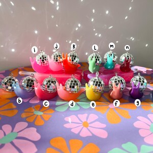May include: A collection of colorful snail-shaped ornaments, each topped with a miniature disco ball. The snails are in various colors, including pink, orange, yellow, green, blue, and purple. They are arranged on a floral patterned surface.