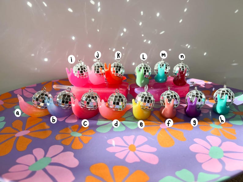 May include: A collection of colorful snail-shaped ornaments, each topped with a miniature disco ball. The snails are in various colors, including pink, orange, yellow, green, blue, and purple. They are arranged on a floral patterned surface.