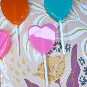 May include: A selection of heart-shaped lollipops in orange, teal, and pink, each on a white stick. The background has a floral pattern in pink, yellow, and purple. The lollipops are a sweet treat.