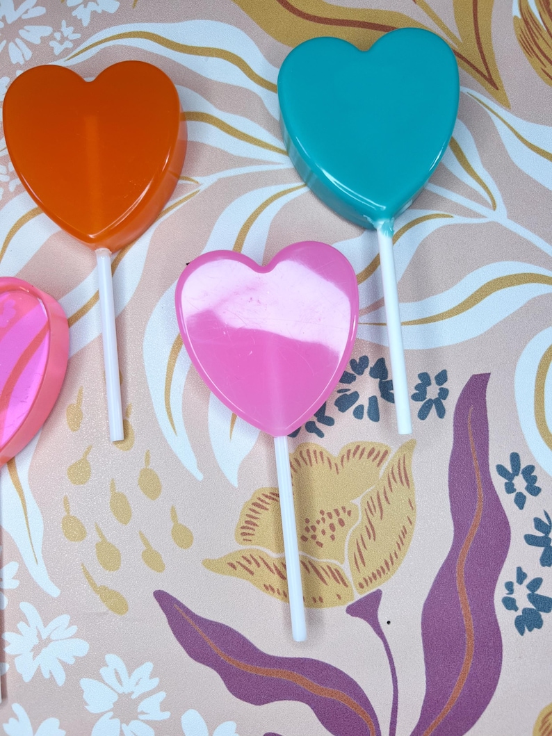 May include: A selection of heart-shaped lollipops in orange, teal, and pink, each on a white stick. The background has a floral pattern in pink, yellow, and purple. The lollipops are a sweet treat.