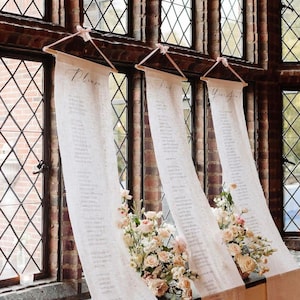 May include: Three white fabric scrolls with calligraphy hanging from a window frame in a brick building. The scrolls are labeled "Please", "Find", and "Your Seat".  The scrolls are decorated with pink ribbons and white flowers.