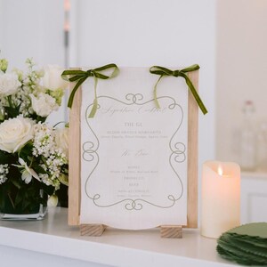 May include: A wooden sign with a white background and green ribbon bows. The sign has a handwritten menu with the title "Signature Cocktails" and lists the drink "The GL Blood Orange Margarita". The sign also lists "Wine, Prosecco, Beer, Non-Alcoholic"