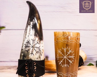 May include: Two drinking horns with intricate carvings. The horn on the left is black and white with a leather strap and a Vegvisir symbol. The horn on the right is brown with a Vegvisir symbol and a decorative band.